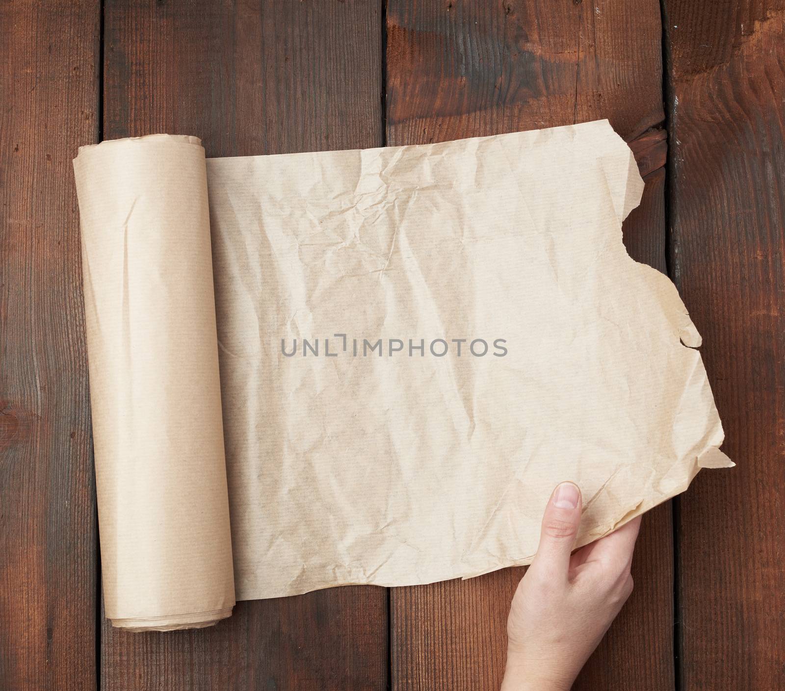hand holds unfolded roll with brown parchment paper on wooden background, top view