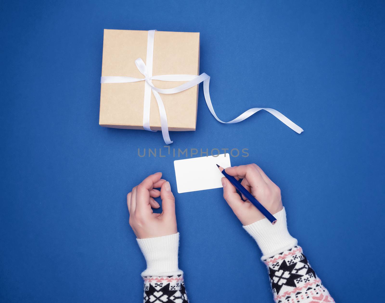 woman holds in her hand a blue wooden pencil and signs a white paper business card, next to a wrapped gift, blue background, top view