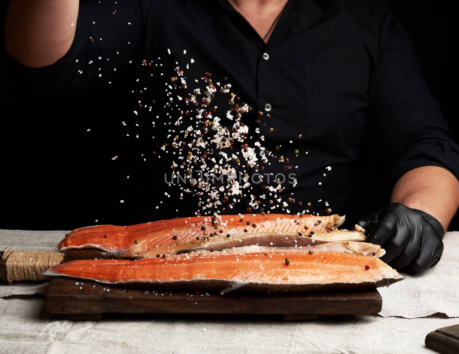 chef in a black shirt and black latex gloves prepares salmon fillet on a wooden cutting board, process of sprinkling with spices and salt, low key