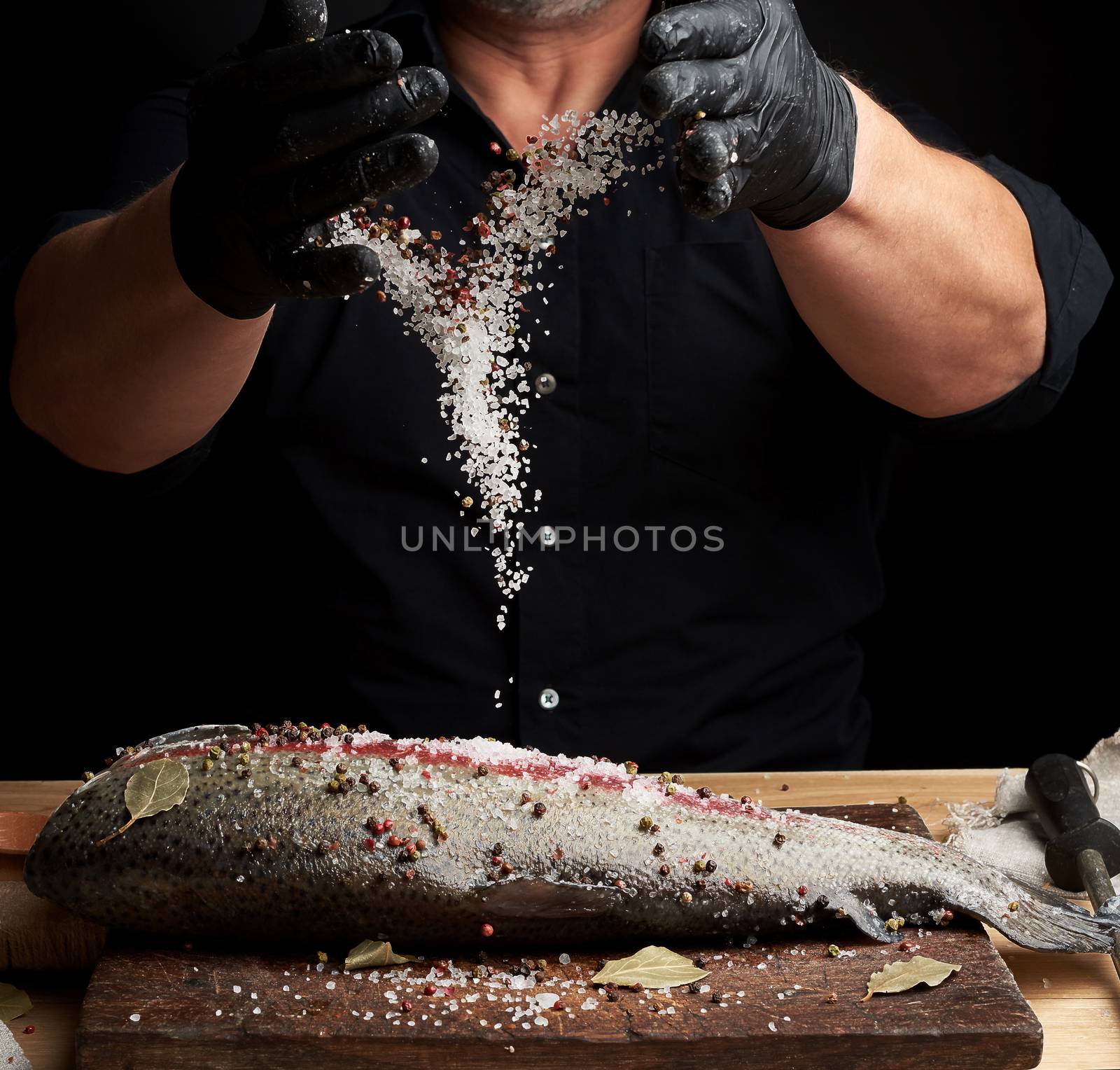 chef in a black shirt and black latex gloves prepares salmon fillet on a wooden cutting board,  process of sprinkling with spices and salt, low key
