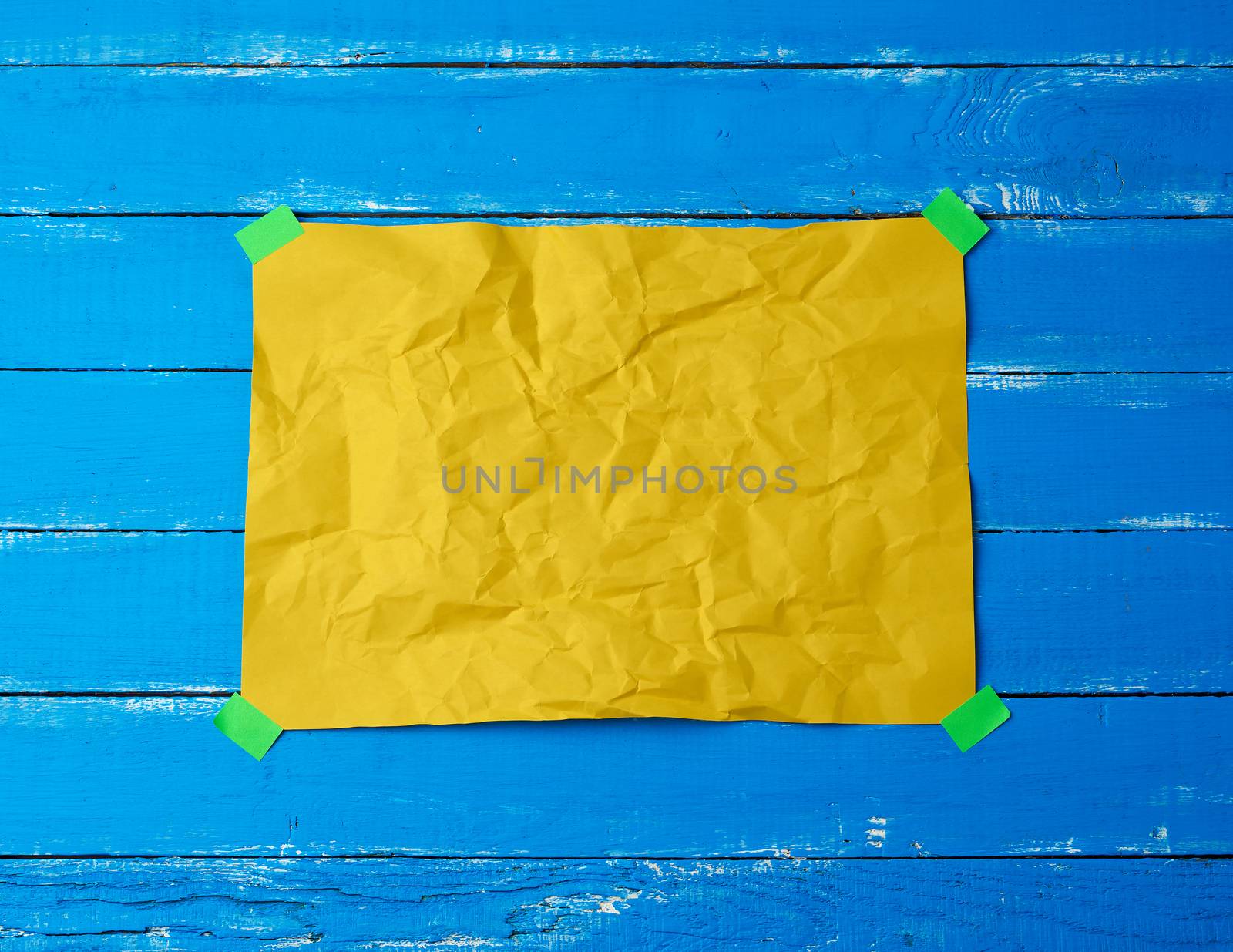 empty yellow crumpled sheet of paper glued with green sticky pieces of paper on a blue wooden background, place for text
