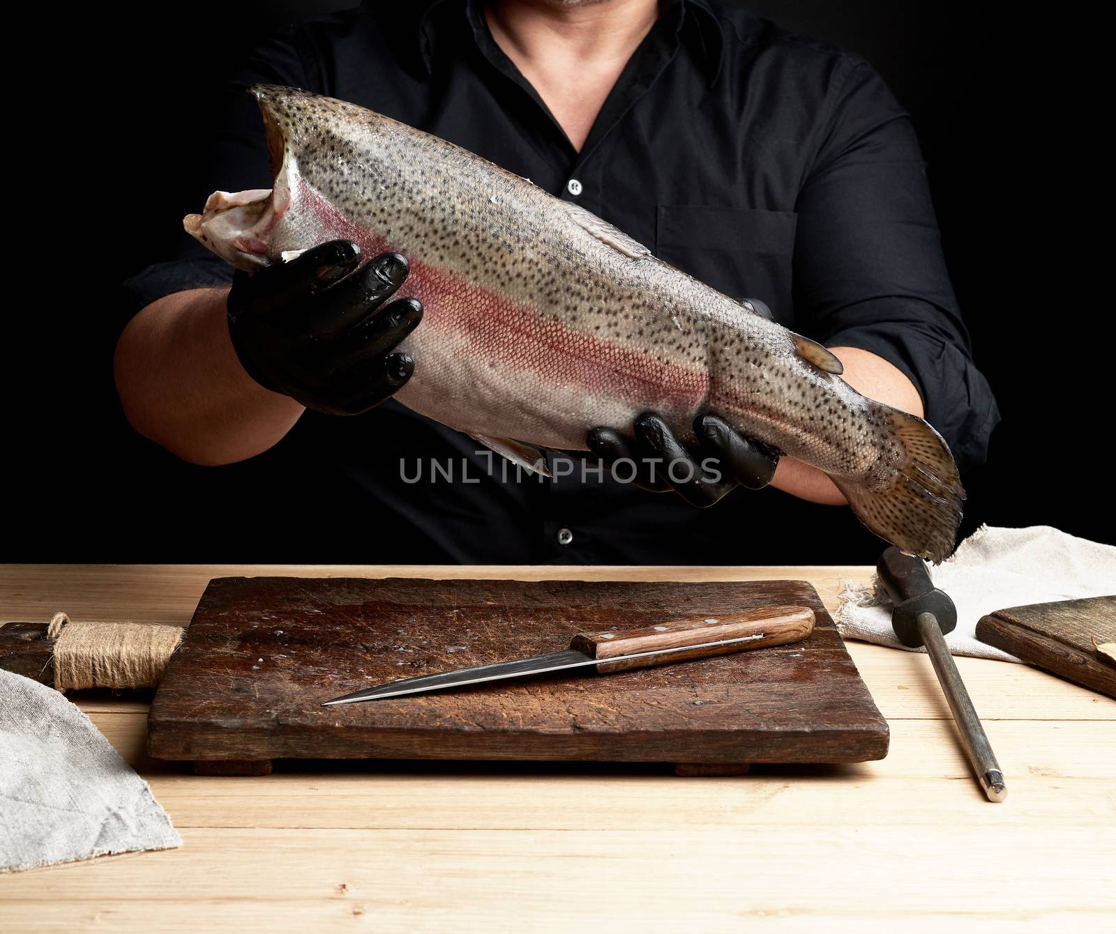 chef in a black shirt and black latex gloves holds a raw carcass of headless salmon fish over a brown wooden cutting board, process of cutting fish