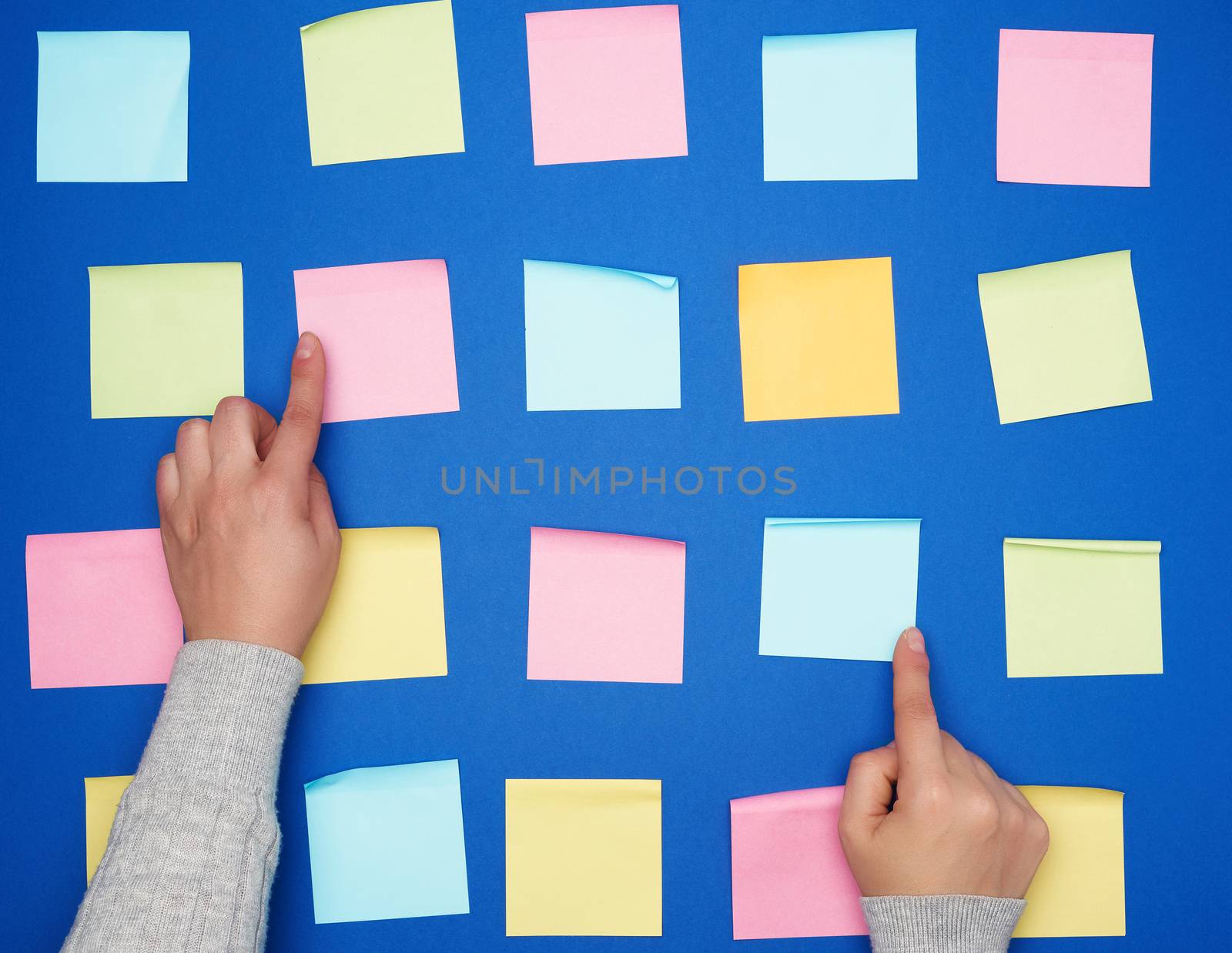 two female hands and a lot of empty paper multi-colored square stickers on a blue background, concept of choosing a strategy with the specified finger