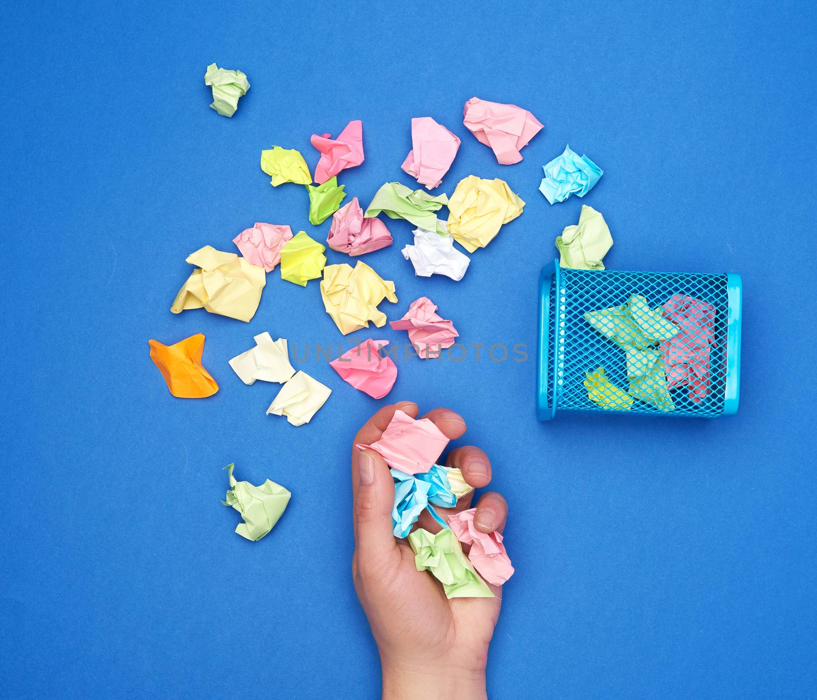 female hand holds a bunch of crumpled multi-colored pieces of paper, next to a metal garbage bin,