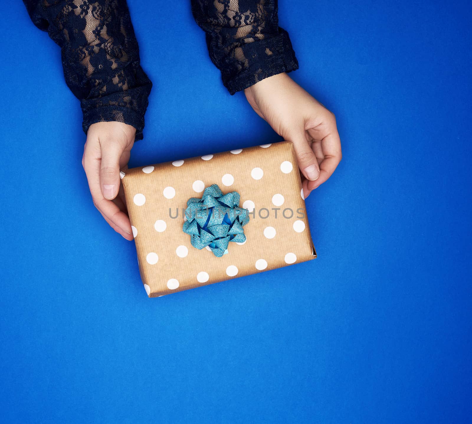 two female hands hold a square brown box with a bow on a blue classic background, top view, festive backdrop
