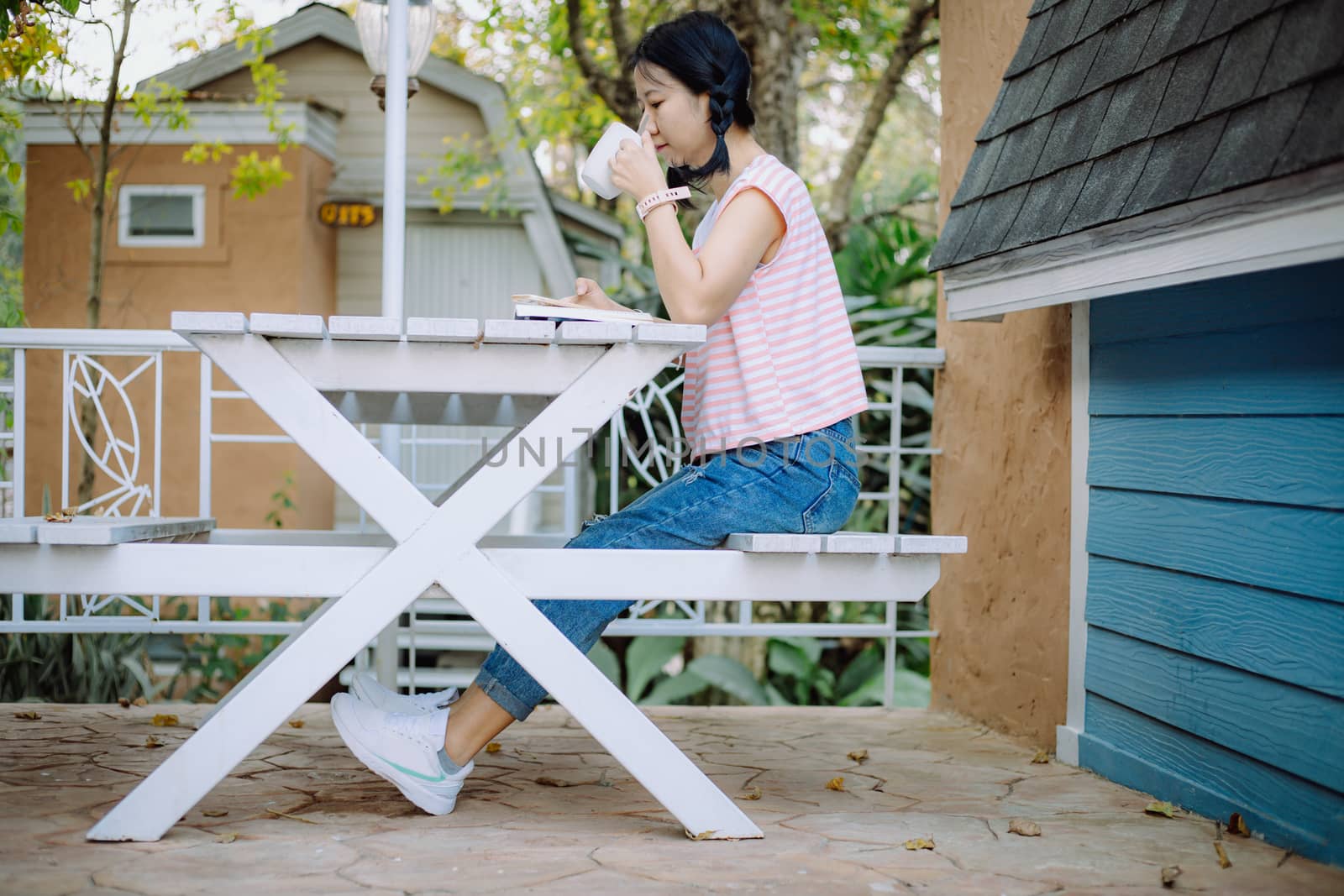 Young asian girl sitting on the white wooden table, drinking a cup of coffee and reading a book at the terraces for relaxation