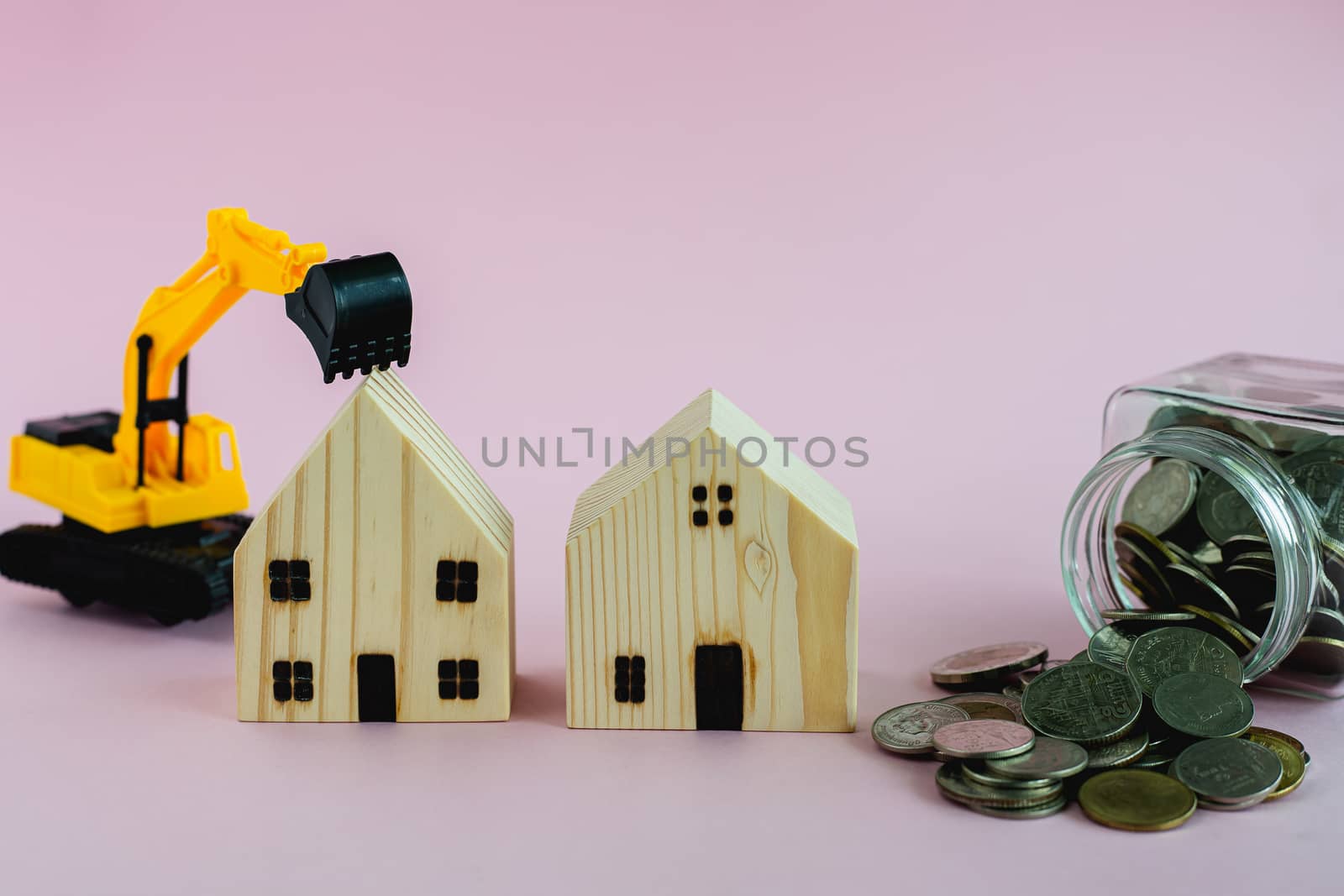 Wooden houses with excavator loader model and money coins in the glass jar on pink background with copy space for business, property finance and construction concept