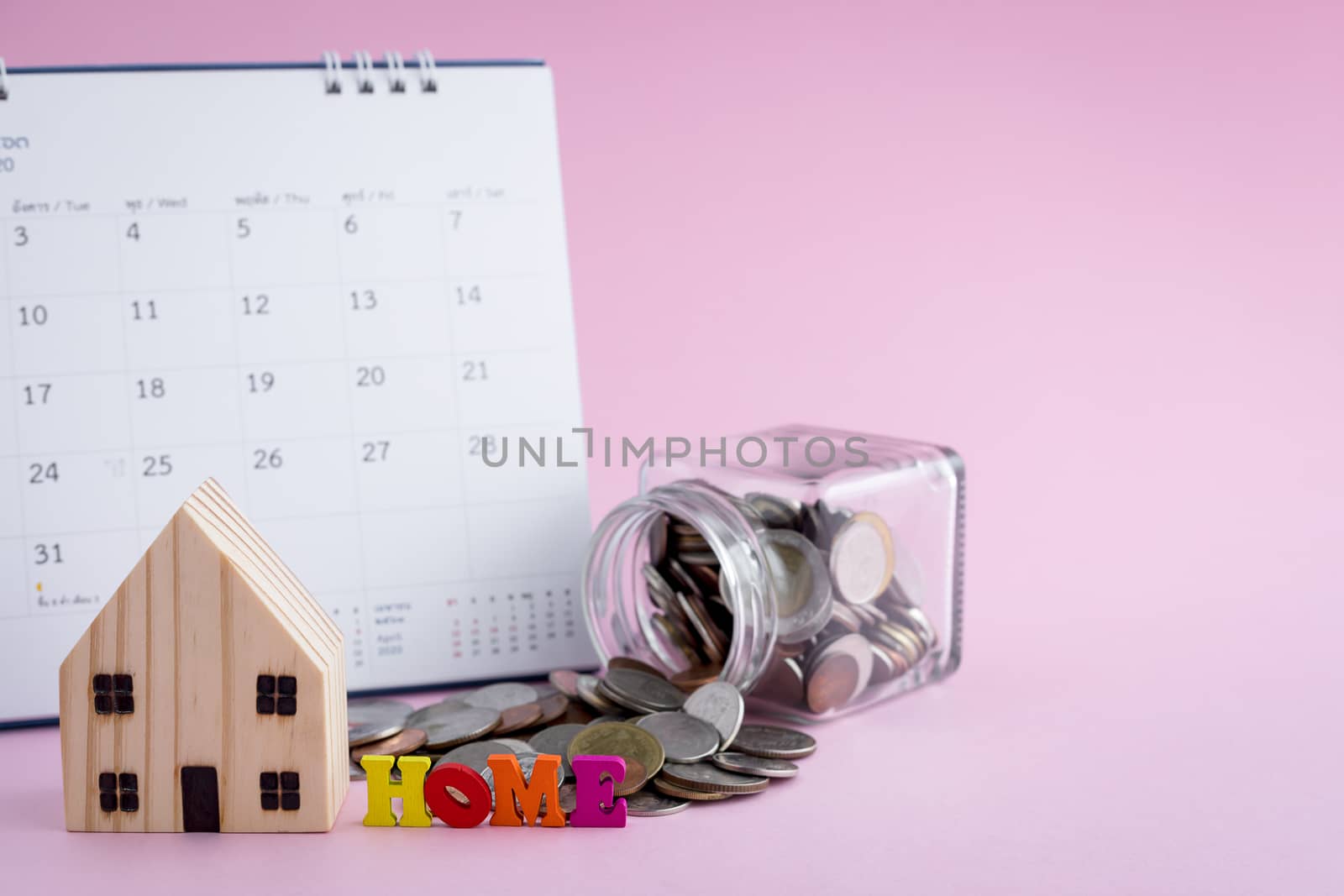 Wooden house model with HOME alphabet, money coins in the glass jar and calendar on pink background for housing business and planning concept