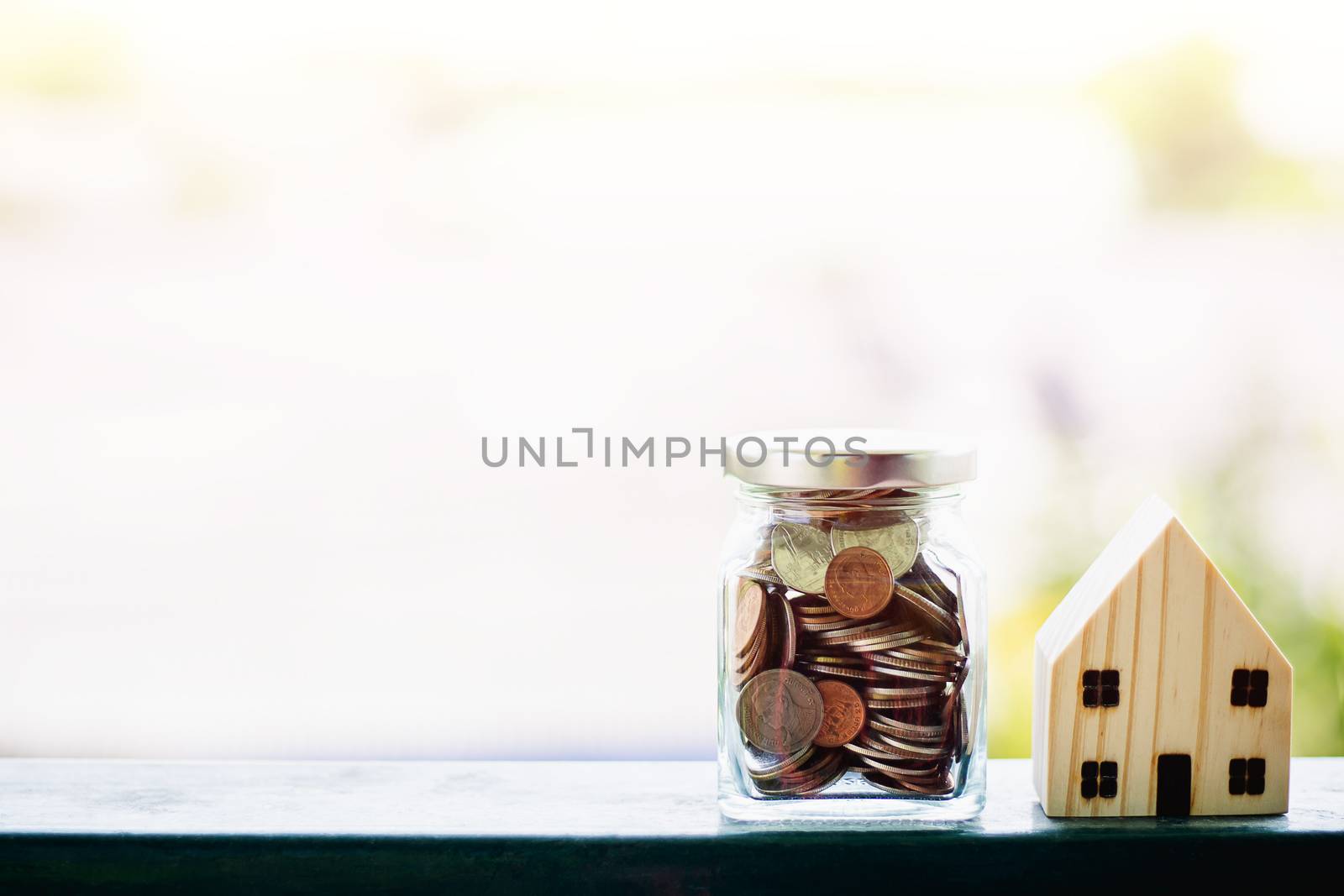 Wooden house model with money coins in the glass jar against blurred natural outdoor background with copy space for business and finance for property concept