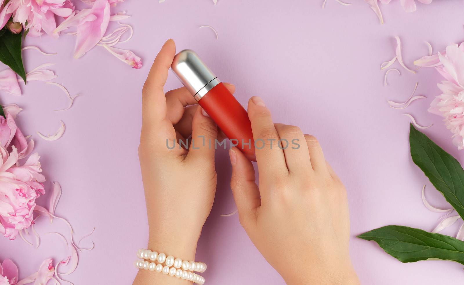 female hands with smooth fair skin keep liquid red lipstick in a tube on a lilac background with pink peonies, top view