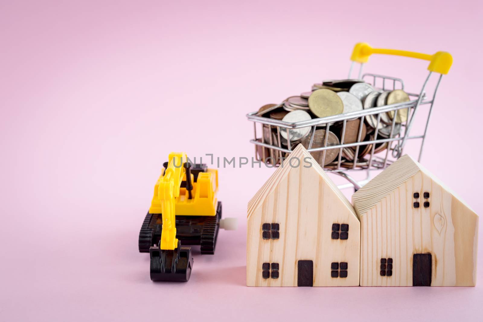 Wooden houses with excavator loader model and money coins in the shopping cart on pink background with copy space for business, property finance and construction concept