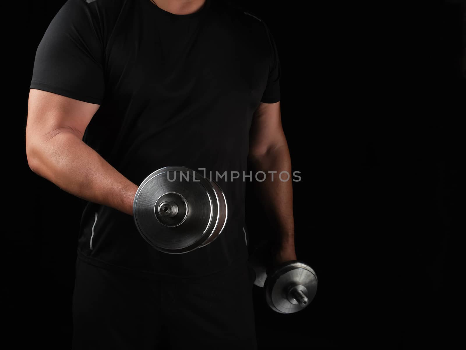 man in black clothes holds steel dumbbells in his hands, his muscles are tense, low key, sports background