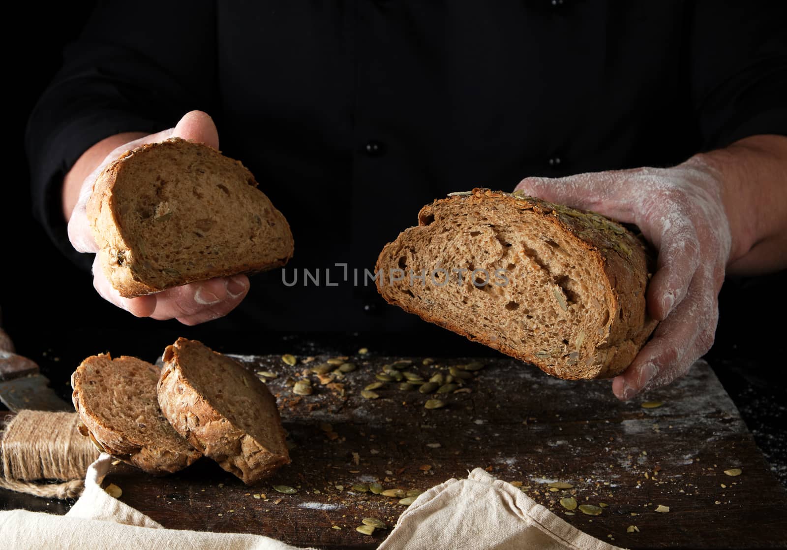 chef in black uniform keeps cut off a piece of bread baked from rye flour and pumpkin seeds, black background
