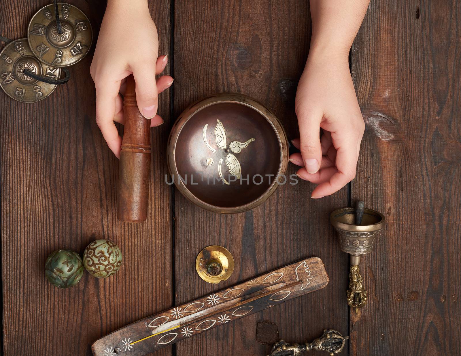 female hands hold a copper Tibetan singing bowl and a wooden stick, on the wooden table are items for alternative medicine, sound massage and meditation