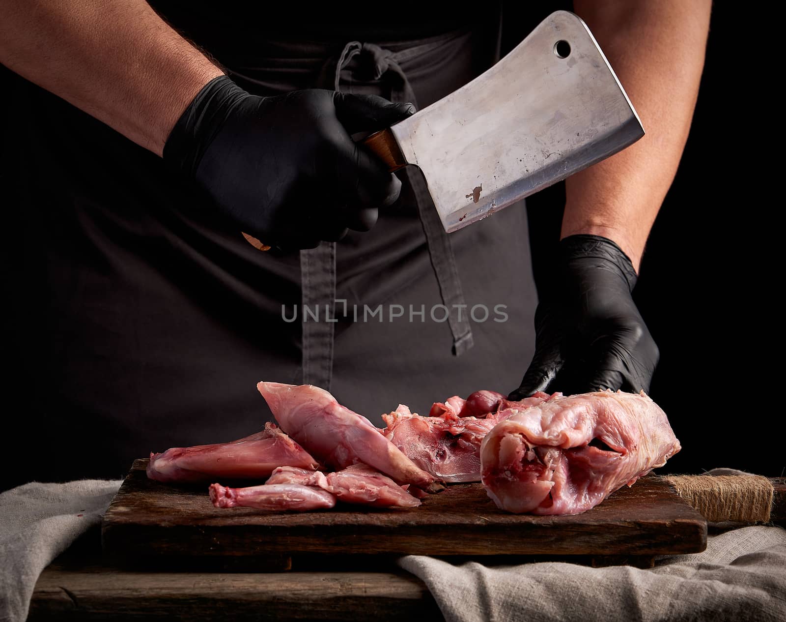 chef in black latex gloves holds a big knife and cuts into pieces raw rabbit meat on a brown wooden cutting board, cooking on a dark background
