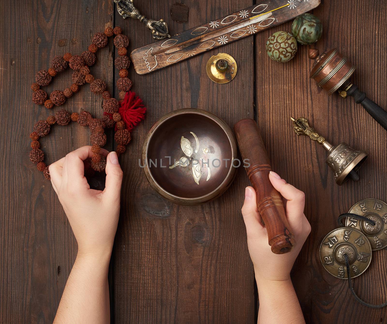 female hands hold a copper Tibetan singing bowl and a wooden stick, on the wooden table are items for alternative medicine, sound massage and meditation