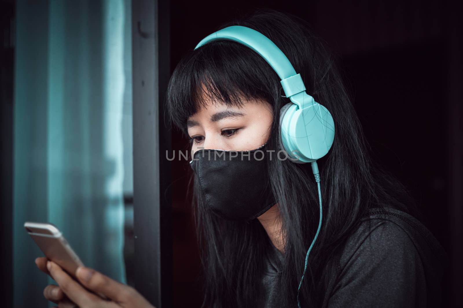Asian woman wearing a black face mask and headphones, using smartphone and staying home for self-quarantine and social distancing in coronavirus or Covid-2019 outbreak situation concept