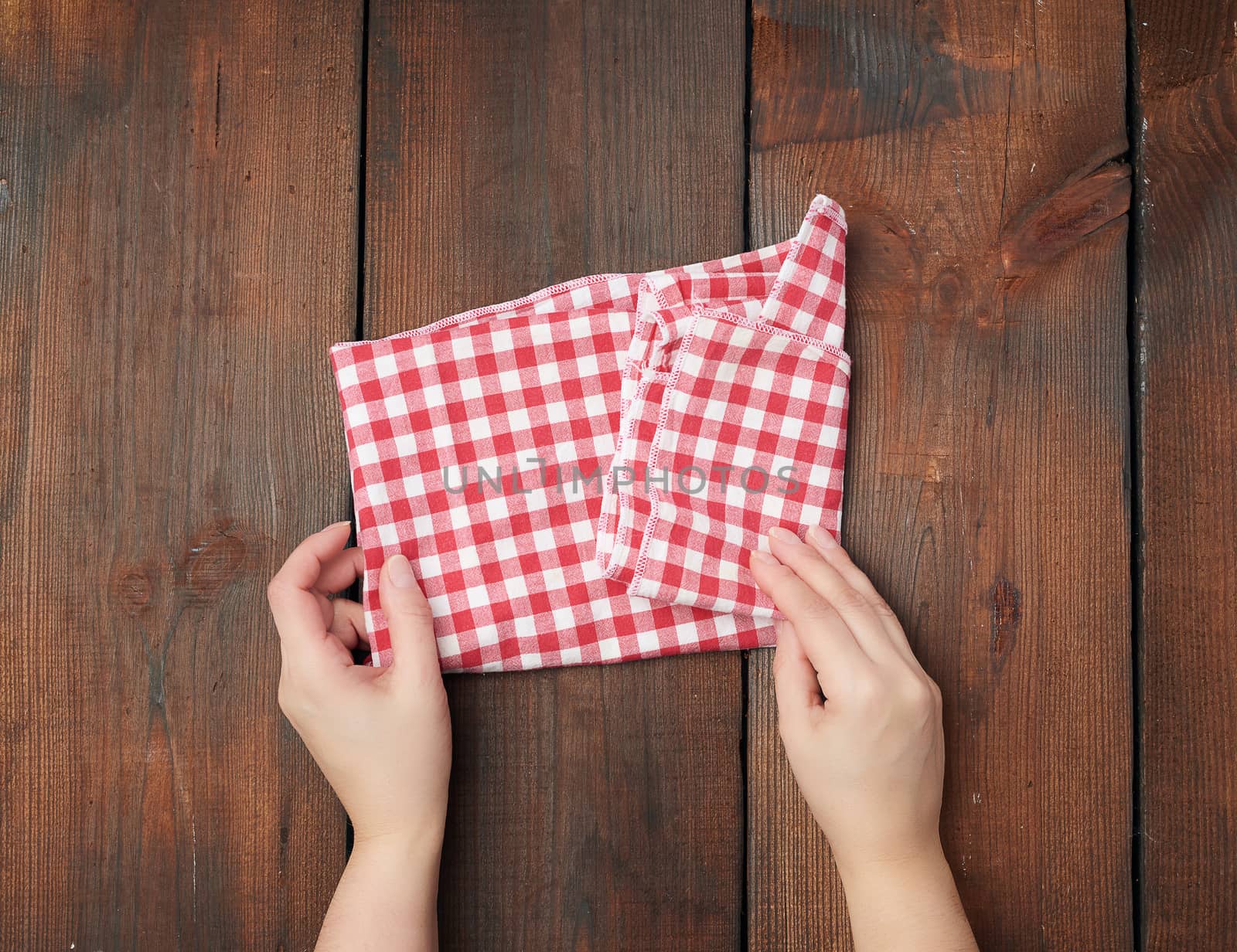 hands hold white red checkered kitchen towel on a brown wooden background, picnic background, top view