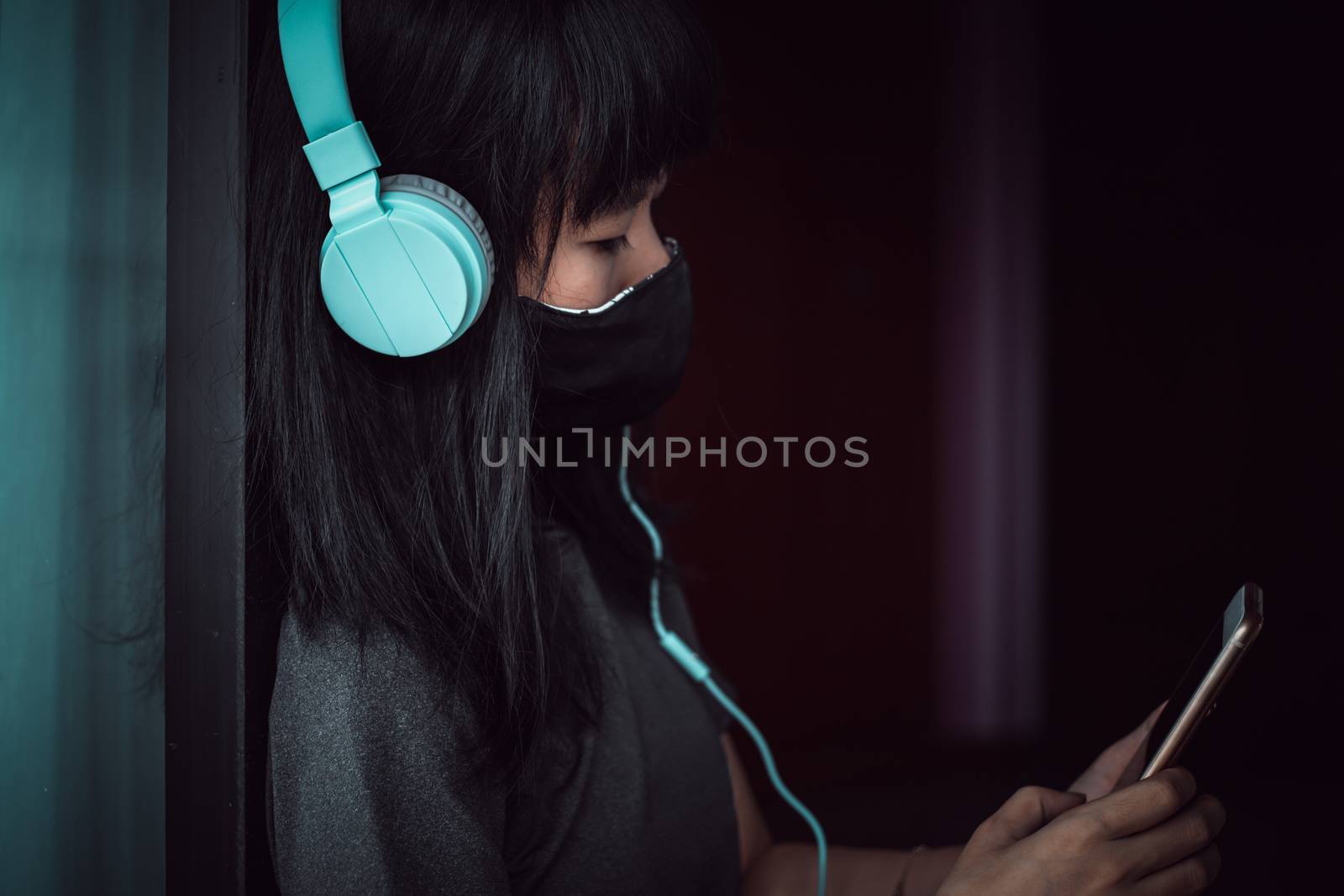 Asian woman wearing a black face mask and headphones, using smartphone and staying home for self-quarantine and social distancing in coronavirus or Covid-2019 outbreak situation concept