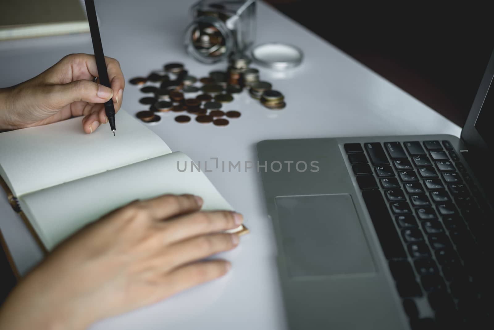 Woman's hand writing a note for planning and saving money with coins in the glass jar and computer laptop for business and finance concept