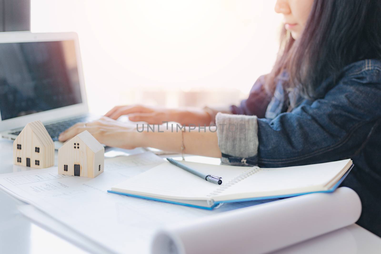 Asian woman using computer laptop with architectural plan and wooden house model on white office desk for building inspection concept