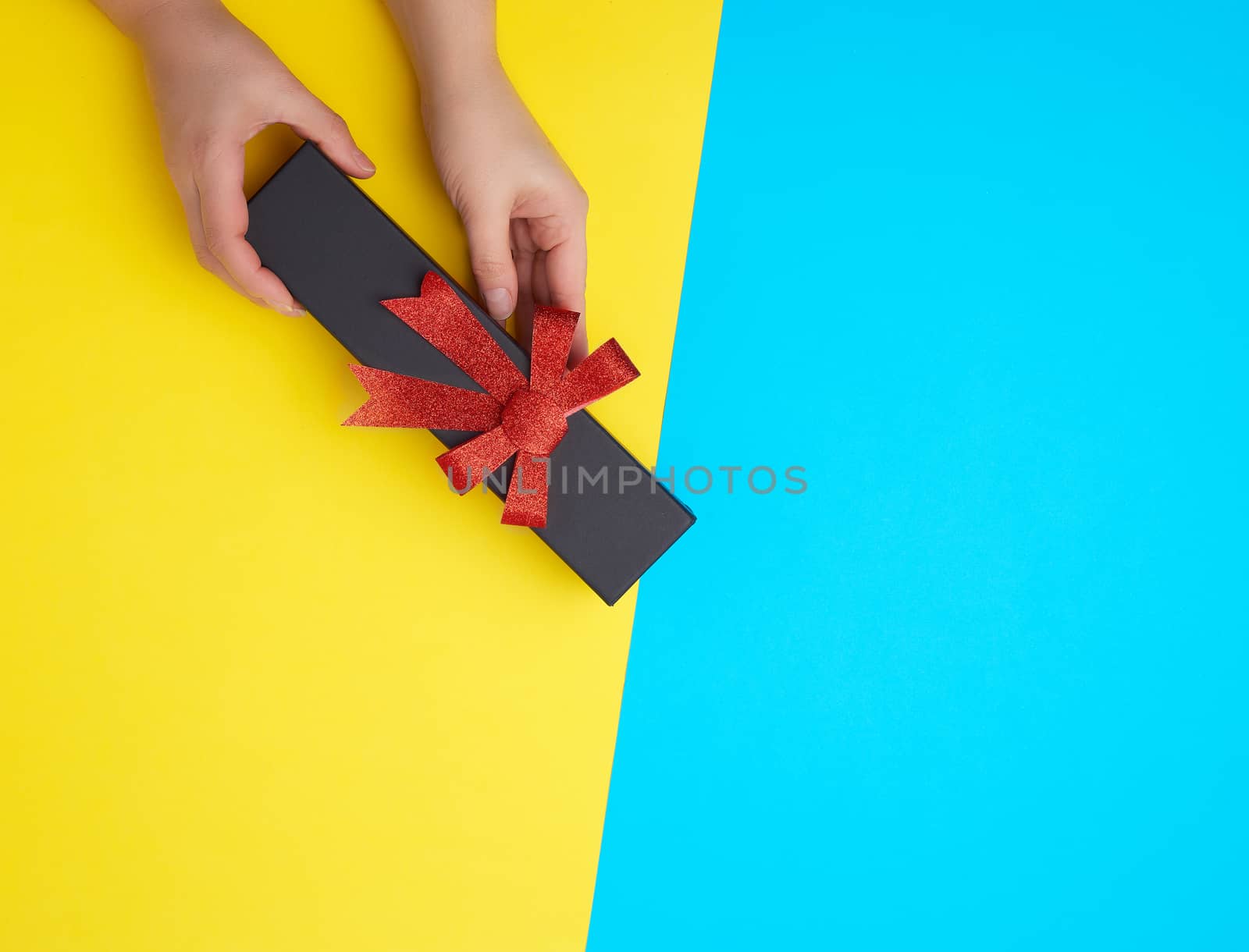 two female hands hold a closed black paper cardboard box with a red bow on a yellow-blue background, concept of surprise, place for text