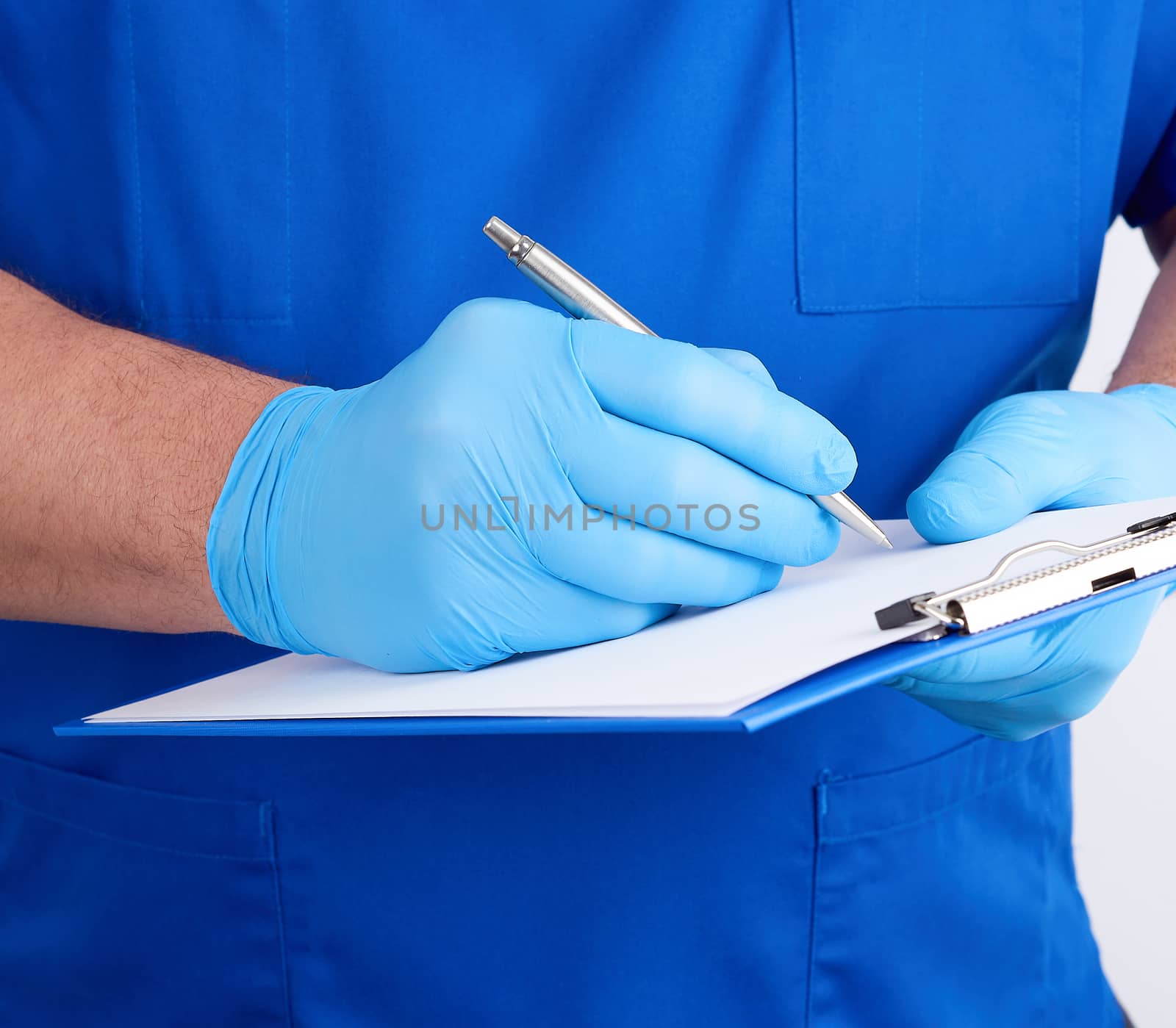 doctor in blue uniform and latex gloves holds a blue holder for sheets of paper and writes with his right hand, white background