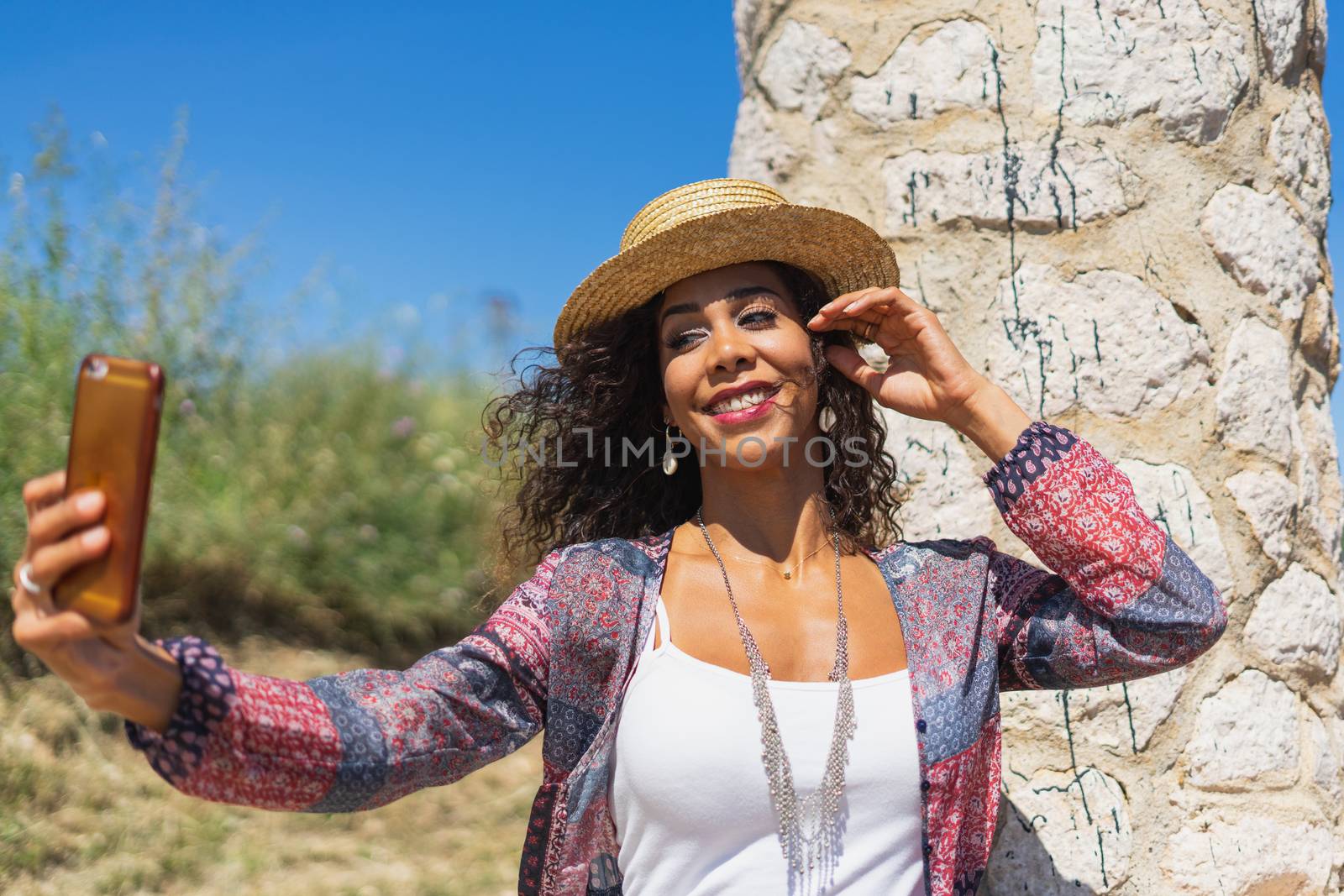 Pretty young Brazilian woman taking a selfie photo with a smartphone outdoors in strong light. Girl smiling expressing energy in good day. Lovely curly woman in stylish hat