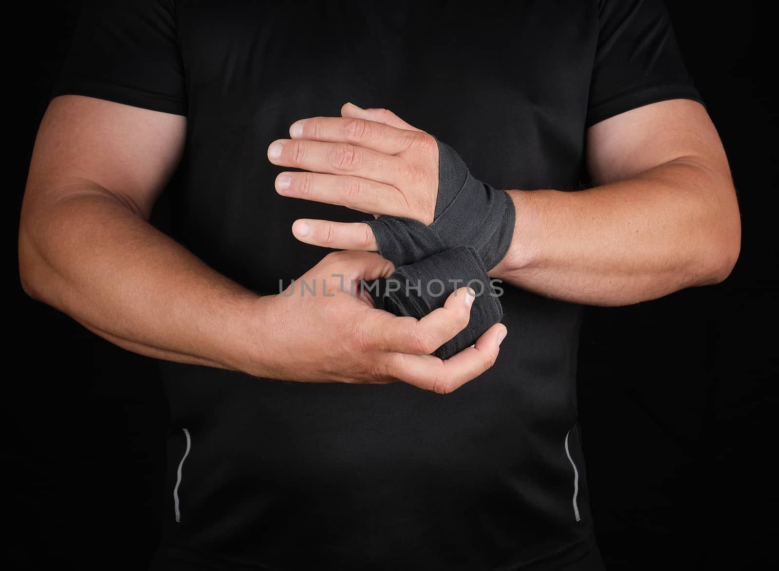 athlete stand in black clothes and wrap his hands in textile elastic bandage before training, dark background
