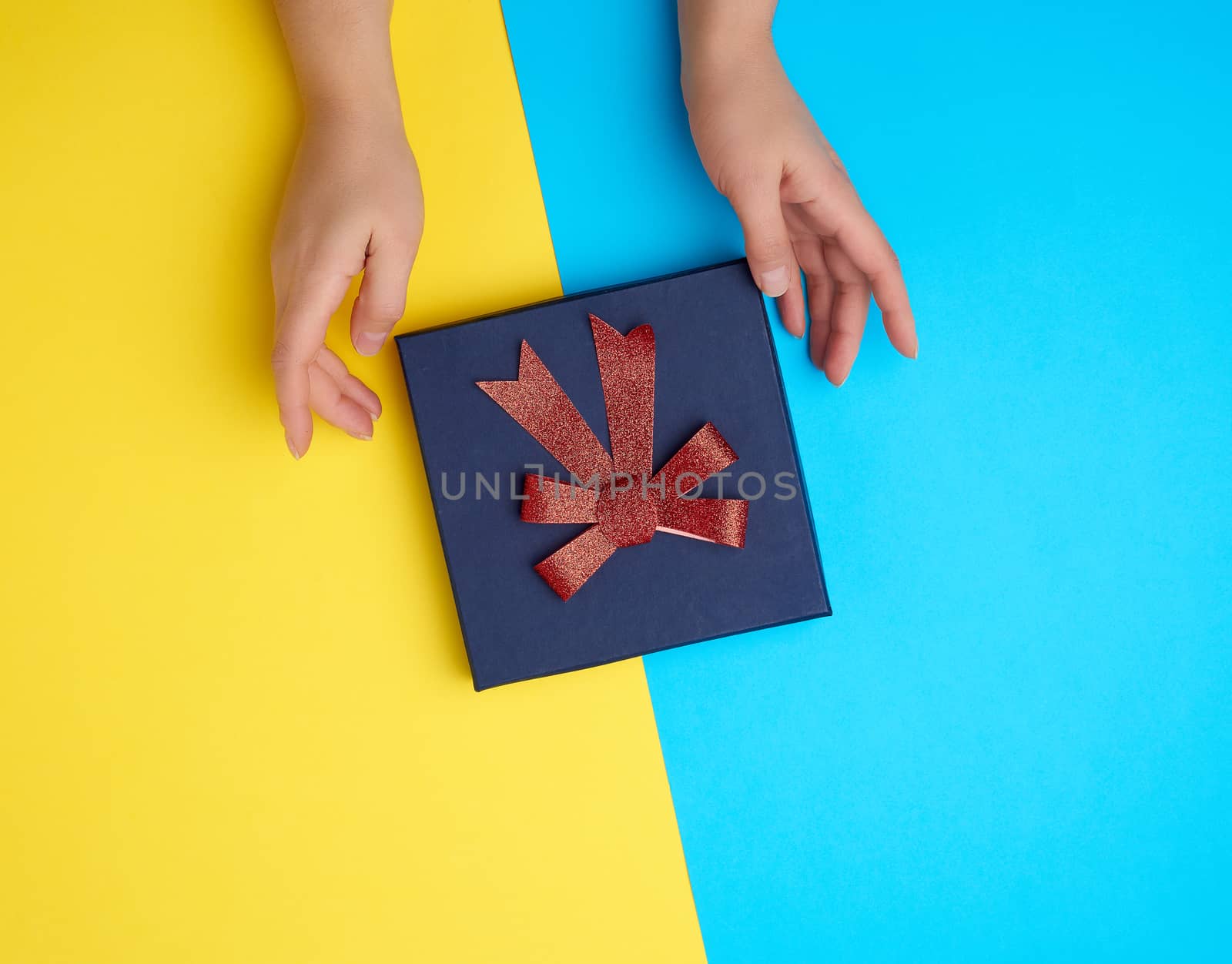 two female hands hold a closed dark blue paper cardboard box with a red bow on a yellow-blue background, concept of surprise, top view