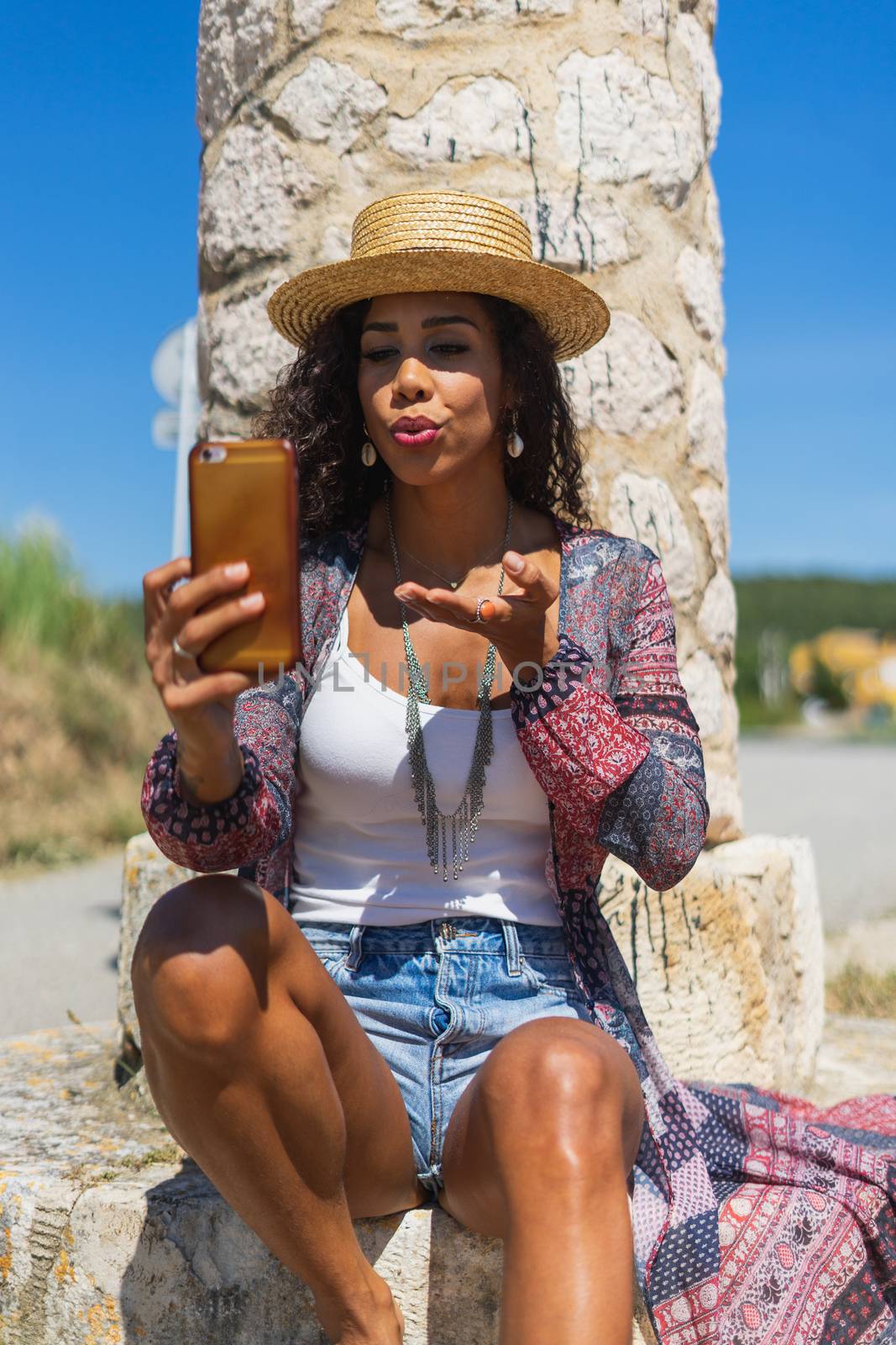 Pretty young Brazilian woman taking a selfie photo with a smartphone outdoors in strong light. Girl smiling expressing energy in good day. Lovely curly woman in stylish hat