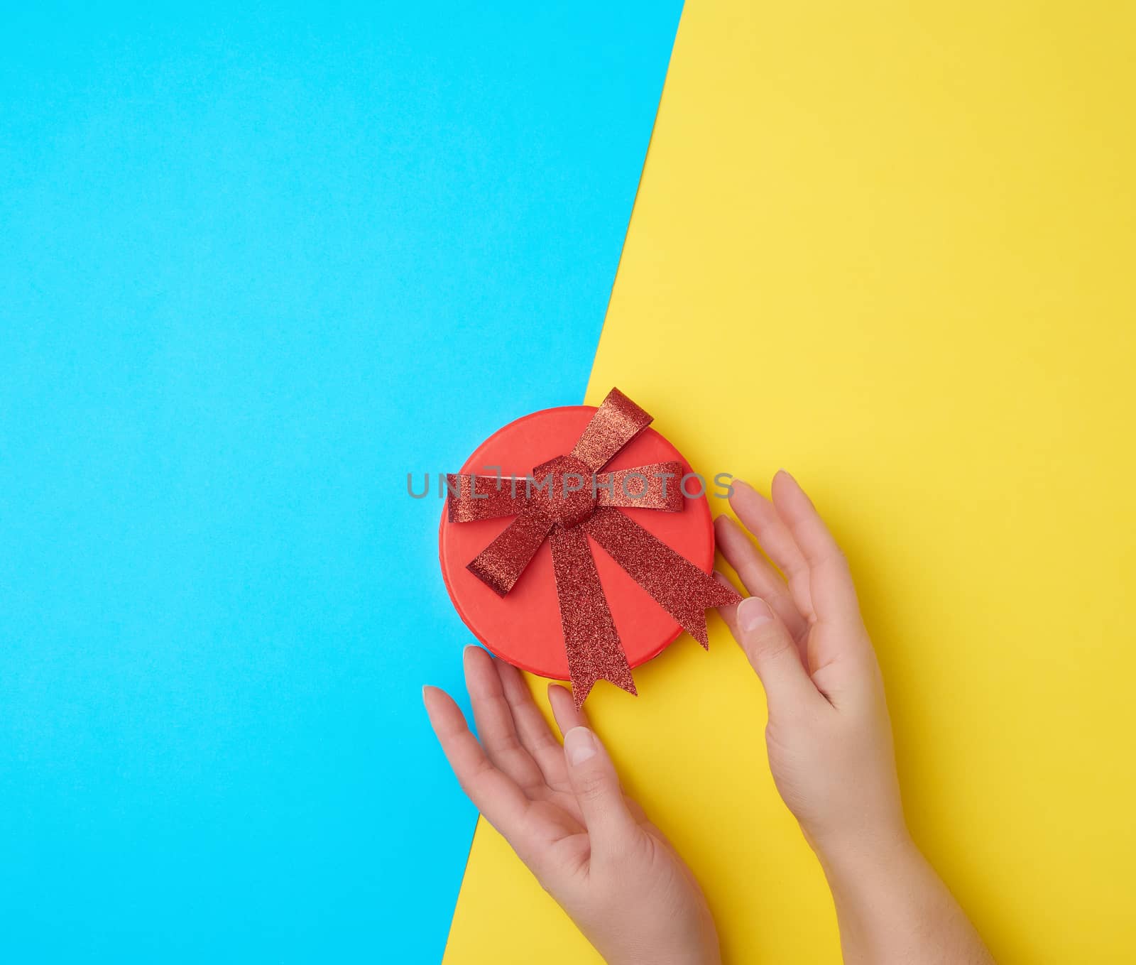 two female hands hold a closed paper cardboard box with a red bow on a yellow-blue background, concept of surprise