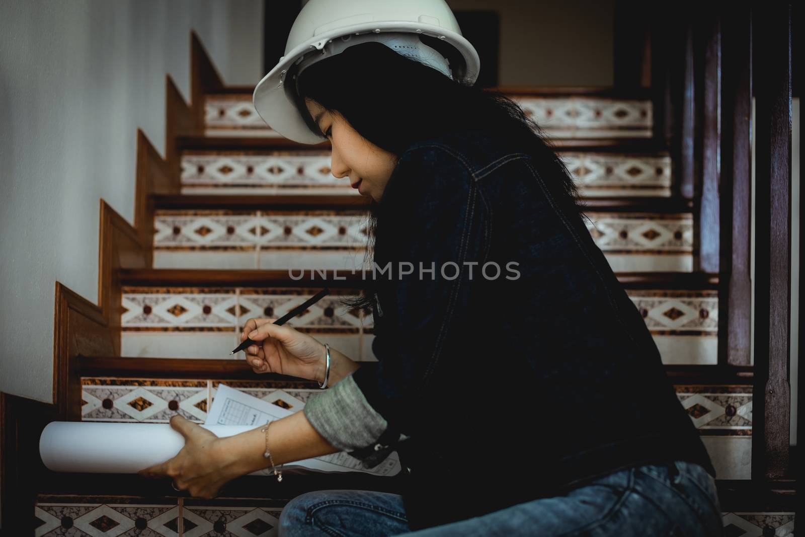 Asian woman looking architectural plan, sitting on the stairs and working at home with safety helmet for building inspection concept