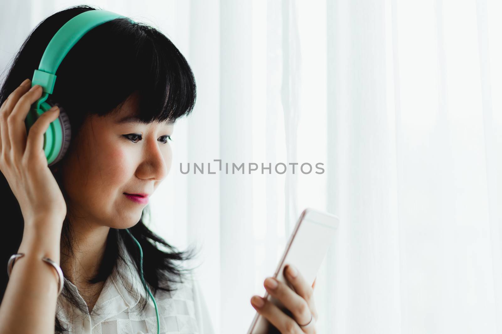 Asian smiling woman wearing headphones, listening music and using smartphone by iamnoonmai