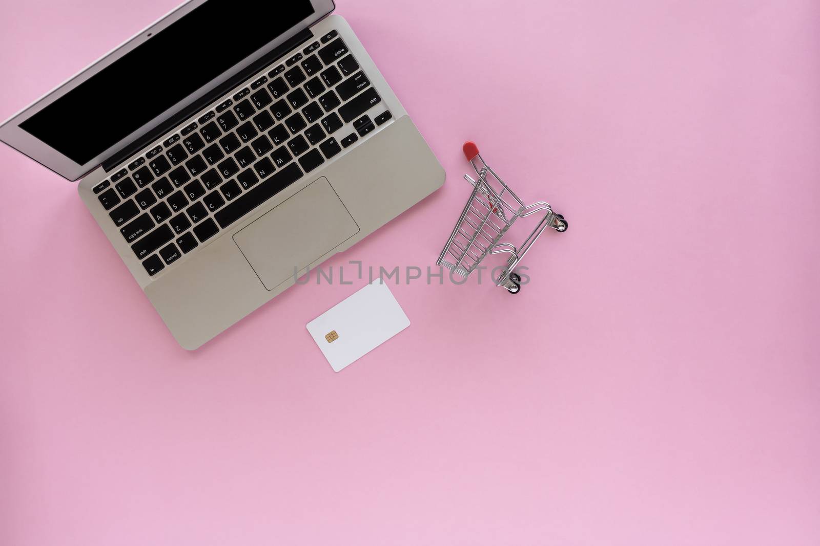 Computer laptop with blank credit card and mini shopping cart or trolley on pink background with copy space for e-commerce, business and finance concept