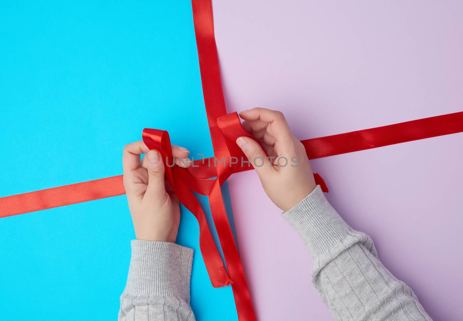 two female hands tie a bow with a satin red ribbon on a gift box, top view