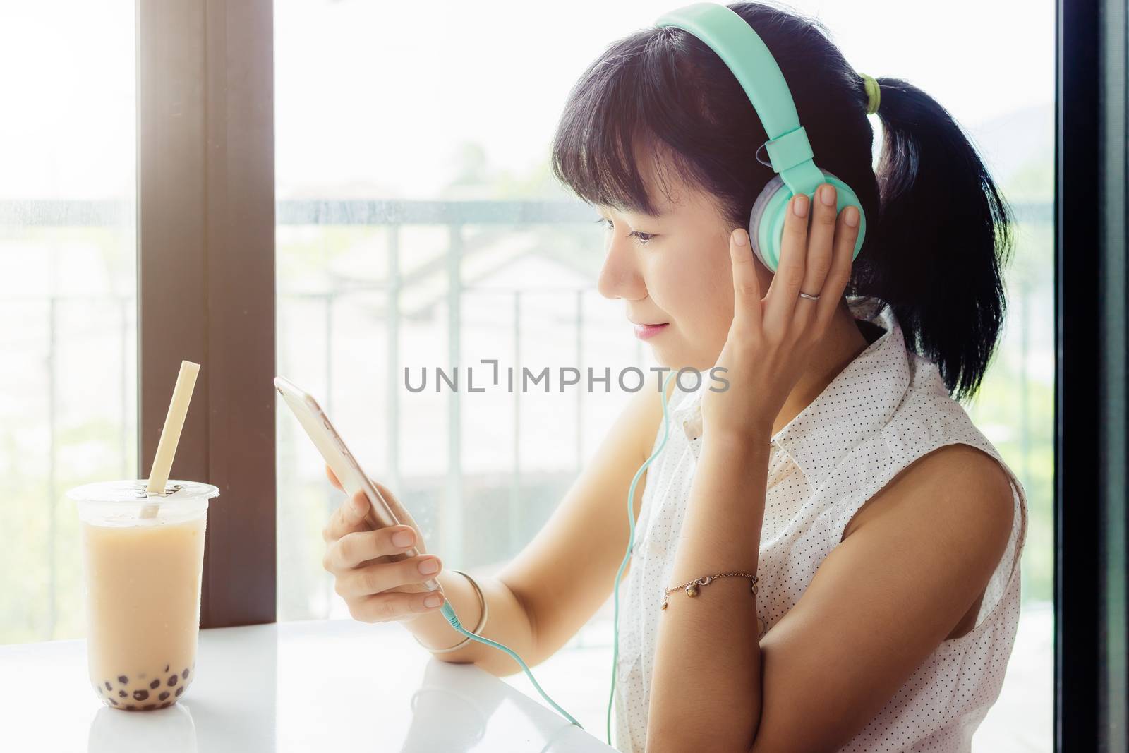 Asian woman wearing headphones and using smartphone with a plastic cup of bubble tea against blurred background for drinks, beverage, staying and working at home concept