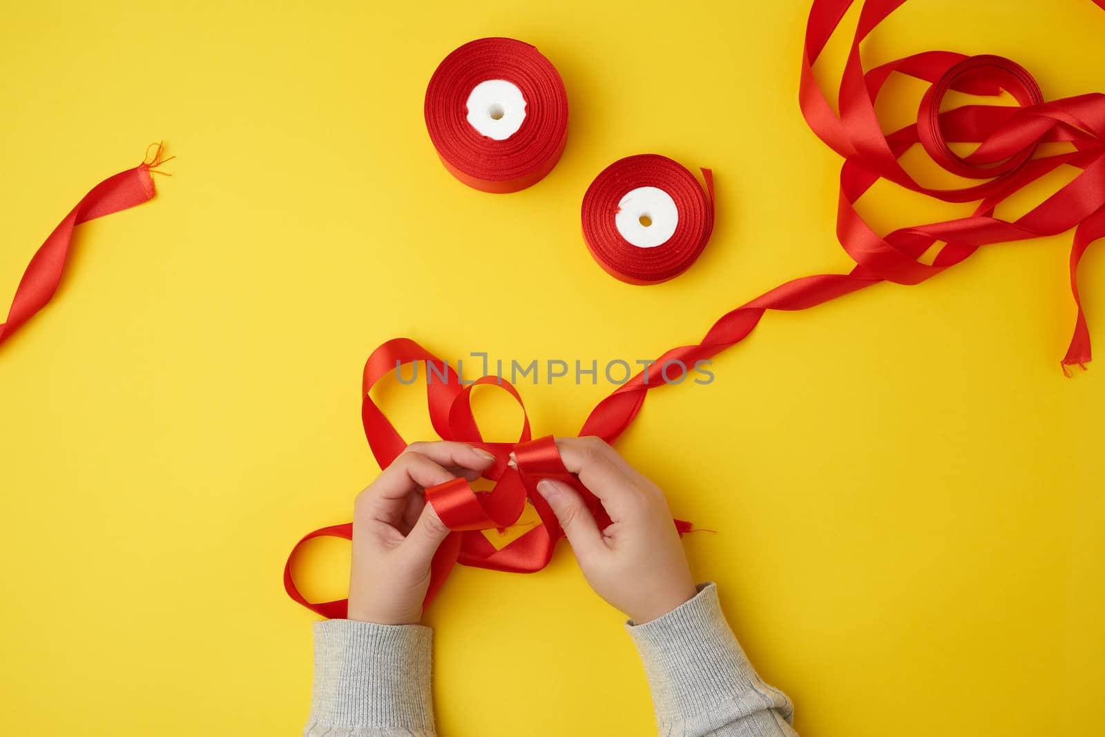 two female hands tie a bow with a red ribbon and reels with a satin ribbon on a yellow background, top view