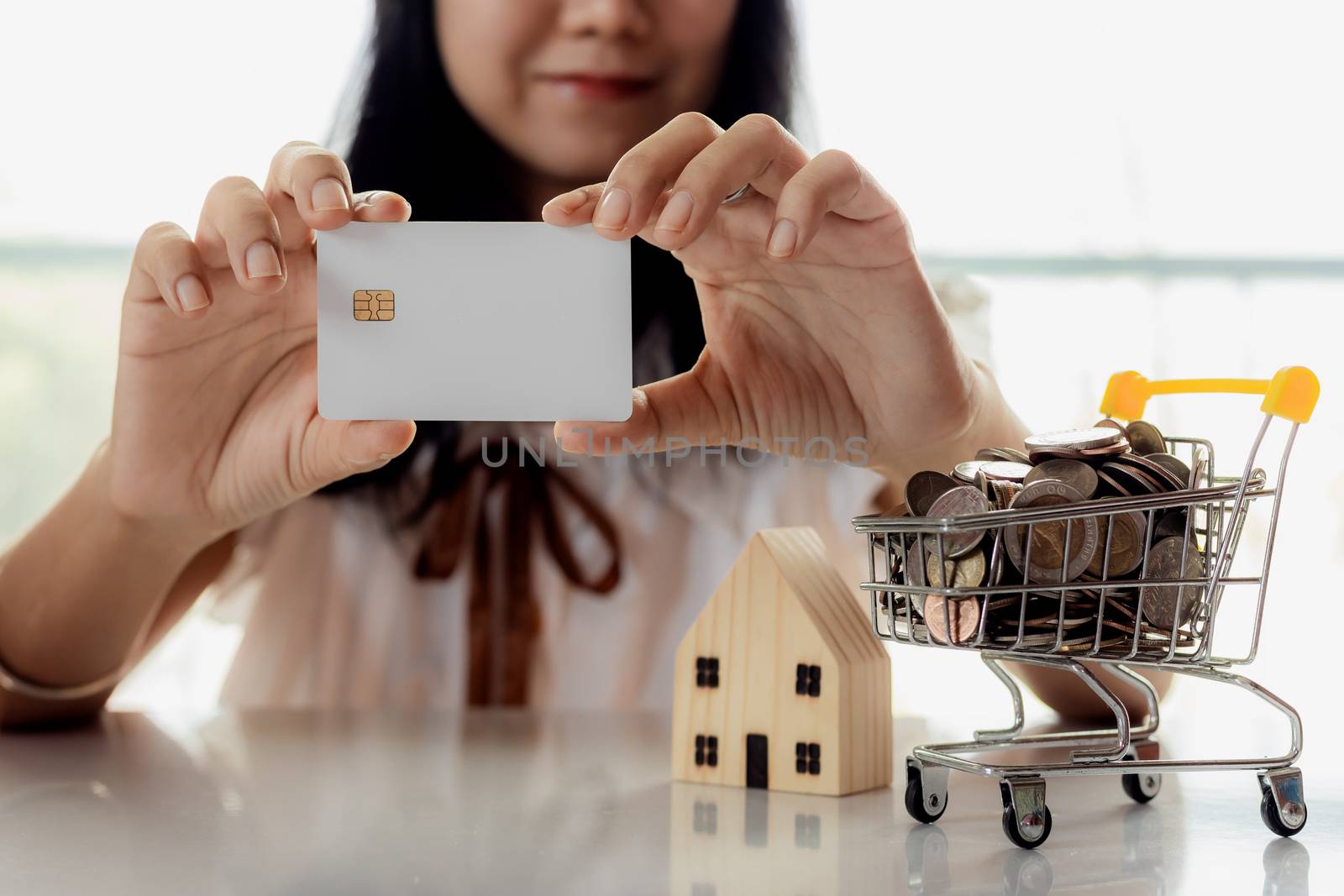 Selective focus of hand holding a blank credit chip card with wooden house model and full of coins in shopping cart for planning, savings and property investment concept