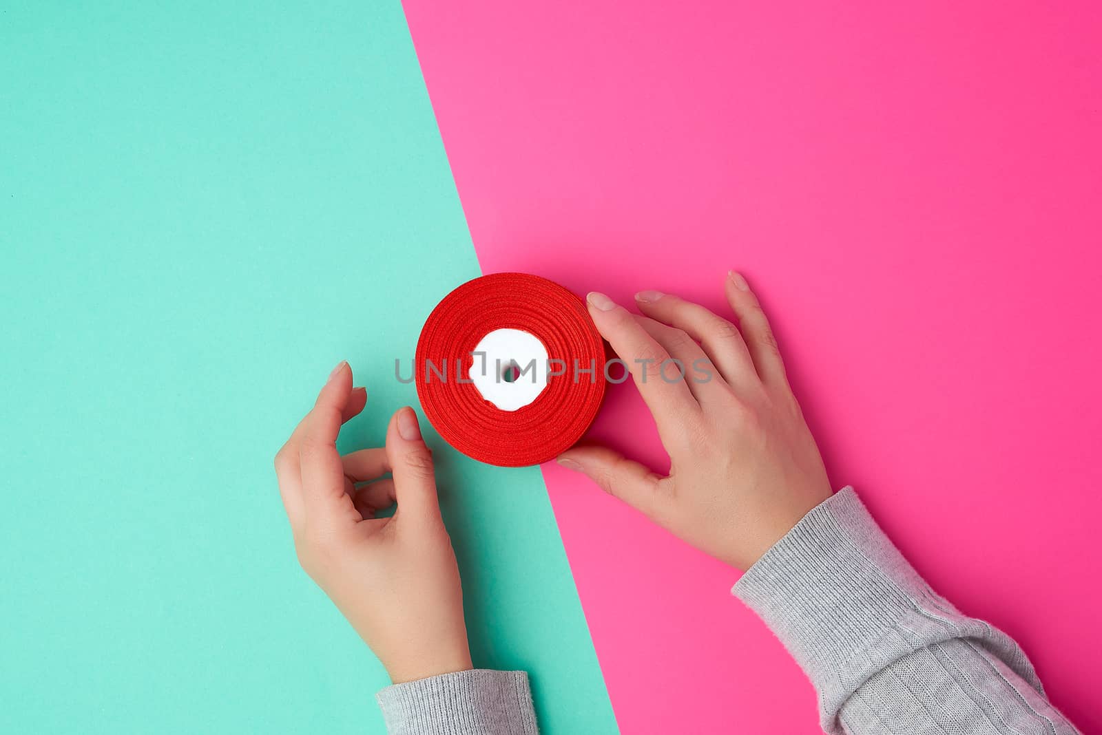 two female hands holding a bobbin with a red silk ribbon on a colored background, top view