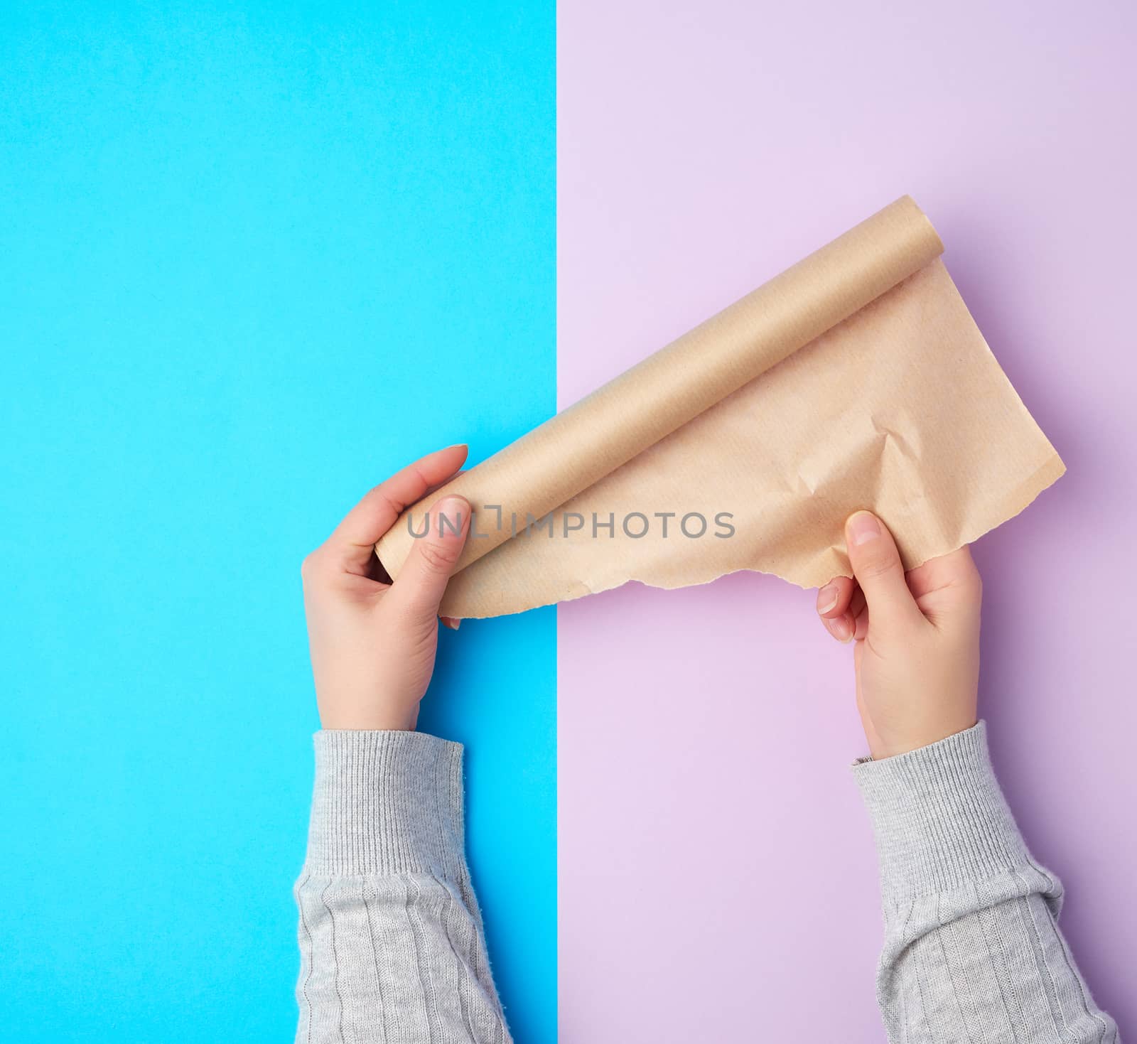 two hands holding a roll of brown parchment paper on a colored background, top view