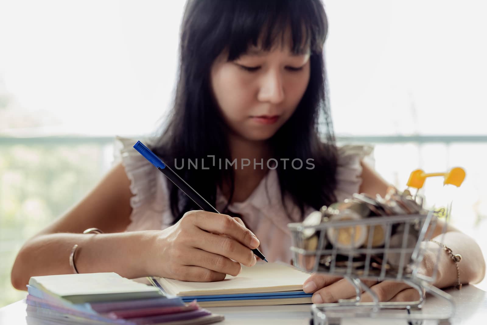 Selective focus of Asian woman's hand writing on a notebook with blurred money and coins in a shopping cart for business and financial planning concept