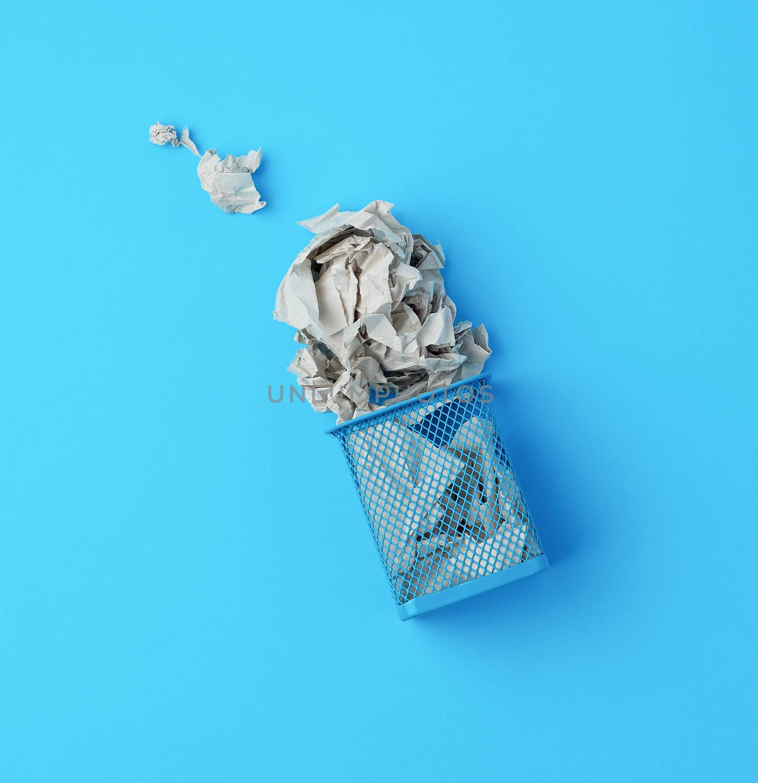 overturned blue metal basket with crumpled pieces of paper, concept of recycling and pollution, blue background