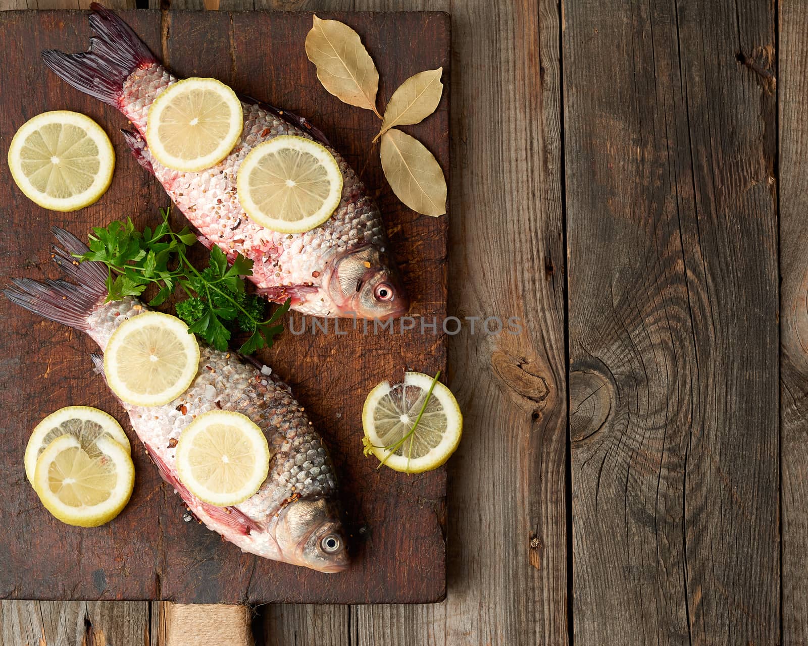 fresh crucian fish sprinkled with spices and lemon slices and lies on a brown wooden cutting board, wooden table from boards, top view