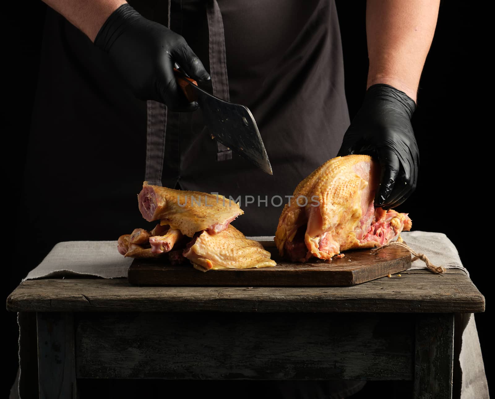 chef in black uniform and latex gloves chopping raw chicken into pieces on a brown wooden board, dark background, cooking process