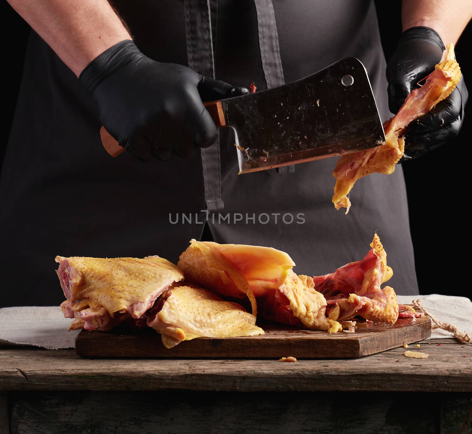 chef in black uniform and latex gloves chopping raw chicken into pieces on a brown wooden board, dark background, cooking process
