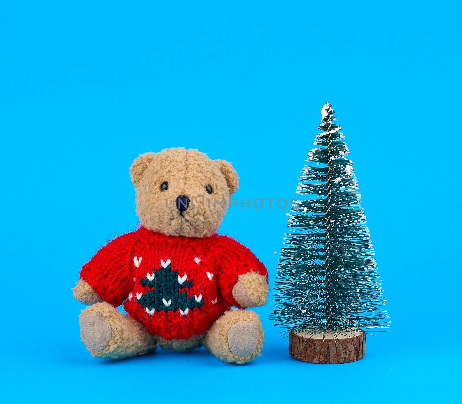 little teddy bear in a red Christmas sweater and a decorative tree on a blue background