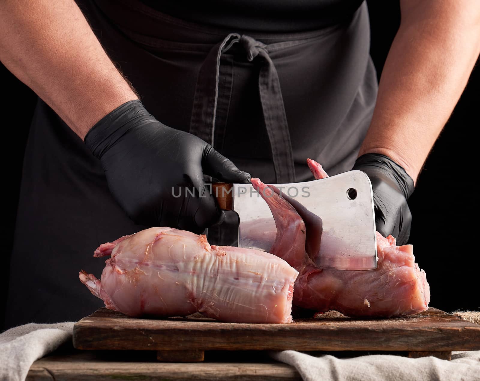 chef in black latex gloves holds a big knife and cuts into pieces raw rabbit meat on a brown wooden cutting board, cooking on a dark background