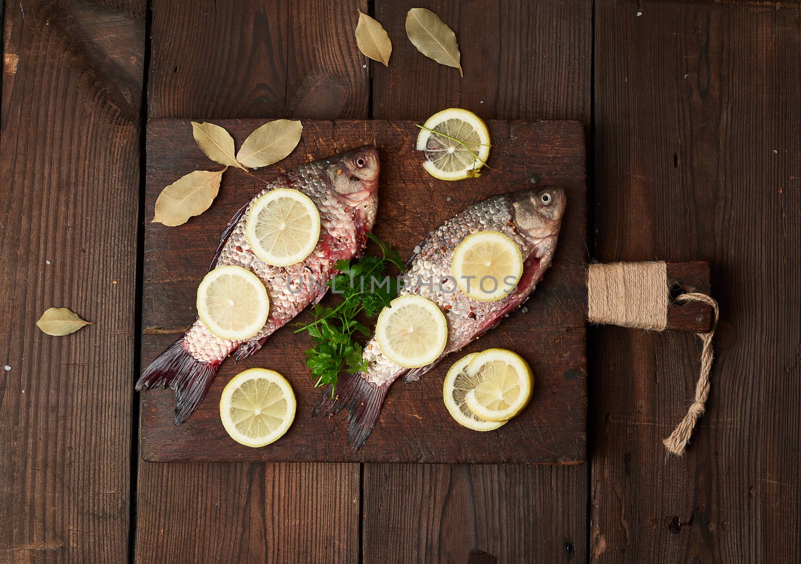 fresh crucian fish sprinkled with spices and lemon slices and lies on a brown wooden cutting board, wooden table from boards, top view