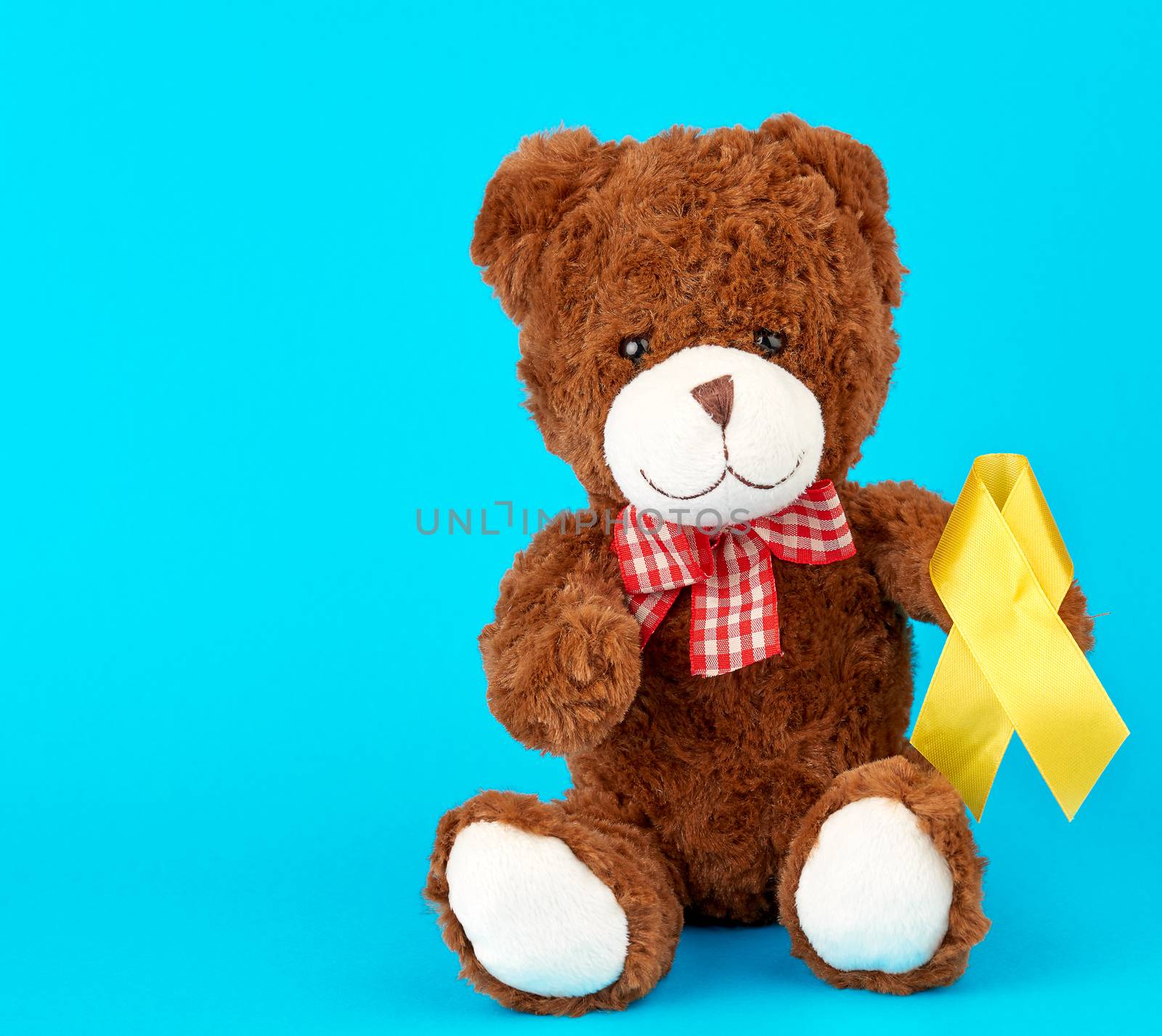 brown teddy bear sits and holds in his paw a yellow silk ribbon on a blue background, concept of the fight against childhood cancer. problem of suicides and their prevention